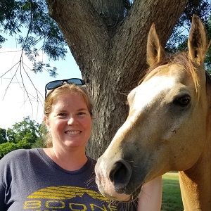 A blonde woman smiles as she poses beside a light-colored horse