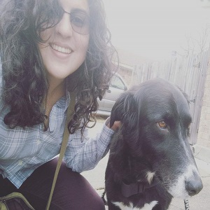 A woman with dark hair and sunglasses smiles at the camera beside a black dog